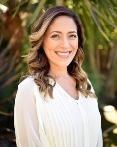 Smiling woman with wavy light brown hair in a white blouse, outdoors with greenery in the background.