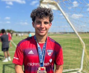 Young male soccer player in a red FC Dallas jersey with a medal around his neck, standing in front of a goal on a sunny field.