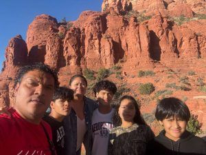 Family of six posing for a selfie in front of orange desert cliffs and a blue sky.