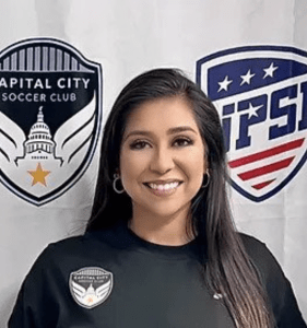 Smiling woman with long dark hair wearing a black shirt poses in front of Capital City Soccer Club and PSL banners with a club badge on her chest.