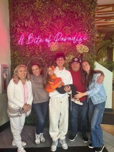 Six people pose together in front of a green plant wall with a pink neon sign reading 'A Bite of Paradise'.