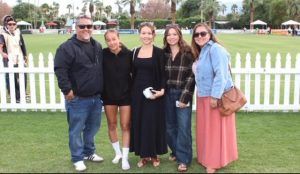 Five people pose for a photo on a grassy field with a white picket fence and palm trees in the background.